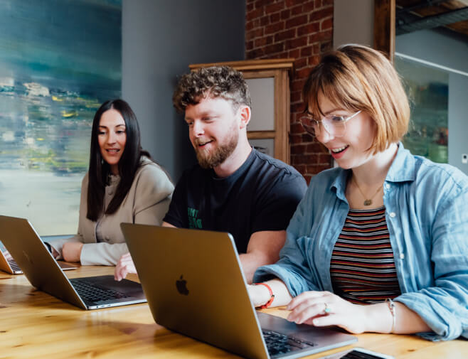  Three people sit at a wooden table using laptops, working together in a casual office environment with modern decor.